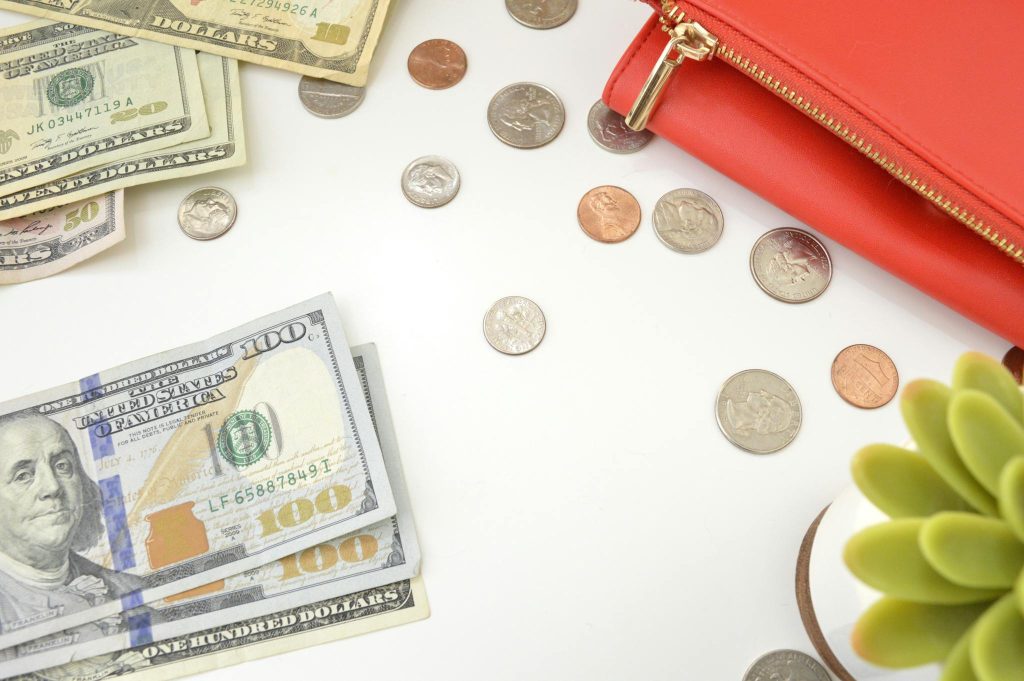 A top view of US dollar bills and coins scattered with a red wallet and succulent plant.