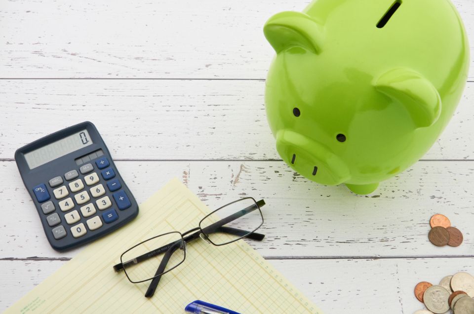 Green piggy bank, coins, calculator and glasses on a desk, illustrating the choice to save or invest